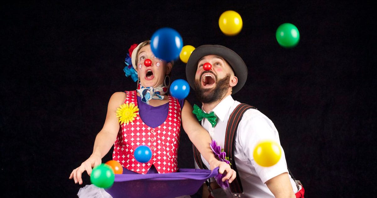A woman dressed in a red vest and a man with a green bowtie both with red noses, laughing at balls flying in the air at the circus